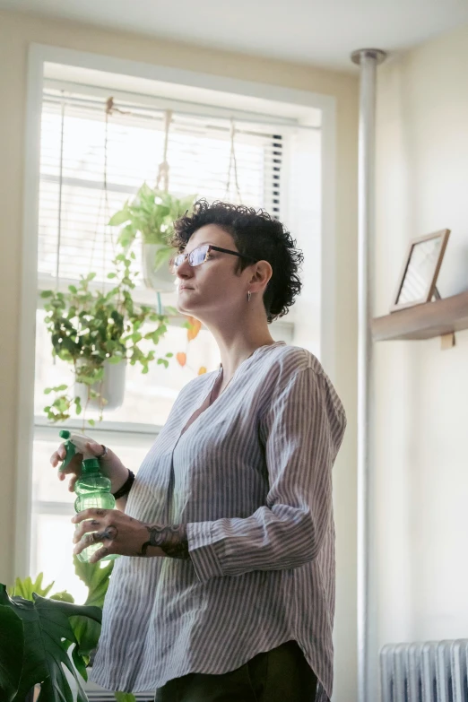 a woman standing in front of a window holding a bottle of water, inspired by Elsa Bleda, plants in glasses, as she looks up at the ceiling, profile image, bright window lit kitchen