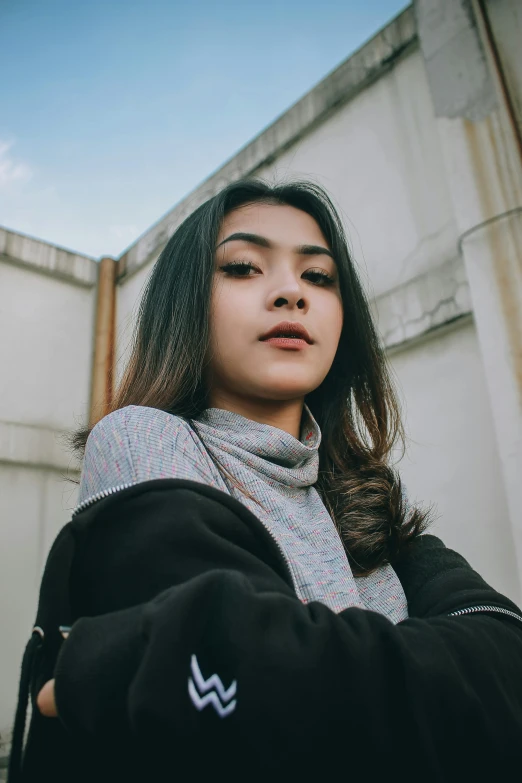 a woman standing in front of a building, trending on pexels, realism, black haired girl wearing hoodie, south east asian with round face, confident pose, contrasted colors