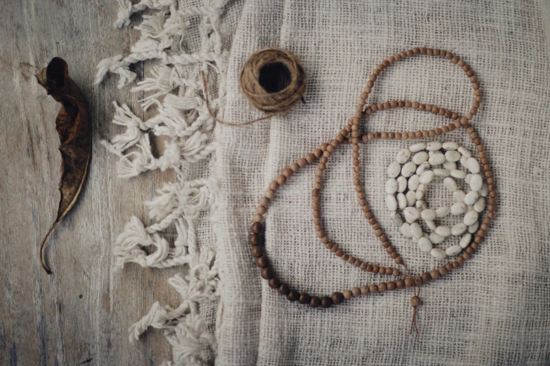 a beaded necklace sitting on top of a table next to a spool of thread, inspired by Marià Fortuny, unsplash, process art, burlap, grain”, buddhist, linen