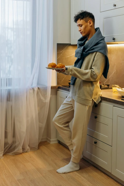 a man standing in a kitchen next to a window, trending on reddit, wearing a baggy pajamas, hijab, holding a baguette, beige color scheme