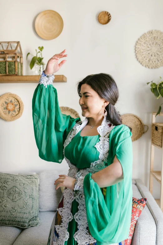 a woman in a green dress standing in a living room, inspired by Li Di, hurufiyya, with arms up, in a white boho style studio, wearing authentic attire, ameera al-taweel