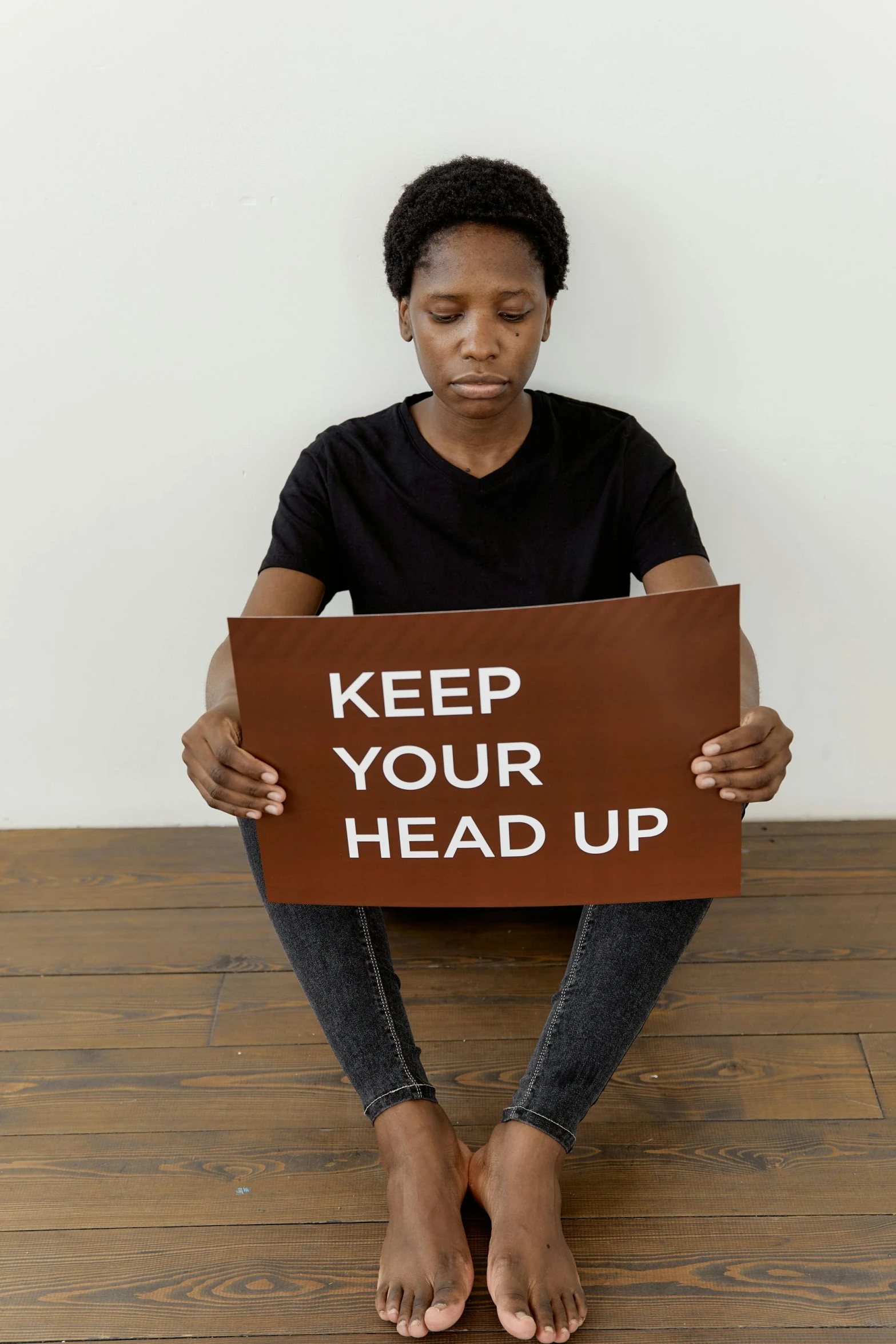 a woman sitting on the floor holding a sign that says keep your head up, by Nina Hamnett, happening, black, large head, panel, brown