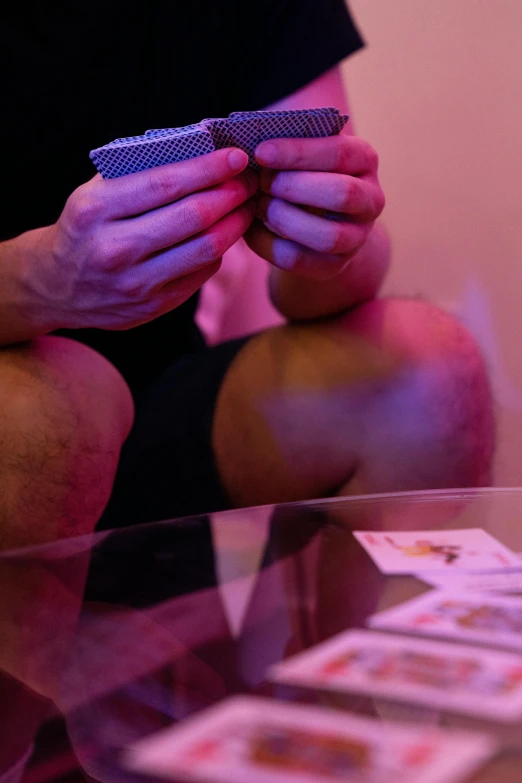 a man sitting on a table holding a deck of cards, purple ambient light, multiple stories, up-close, playing games