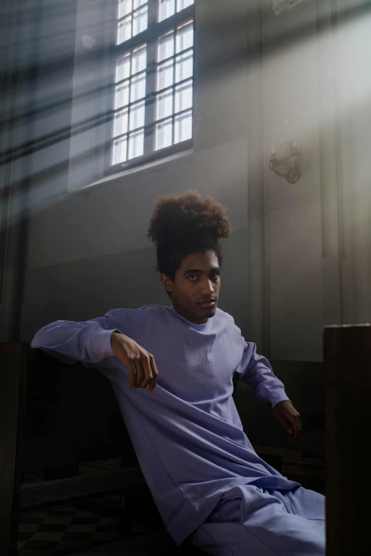 a man sitting on a bench in front of a window, an album cover, by Jacob Toorenvliet, pexels contest winner, renaissance, black teenage boy, portrait androgynous girl, wearing a purple sweatsuit, in church