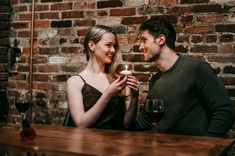 a man and woman holding wine glasses in front of a brick wall, by Lee Loughridge, pexels contest winner, eating cakes, handsome girl, smiling at each other, sitting on a mocha-colored table