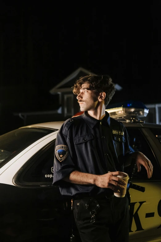 a police officer standing next to a police car, an album cover, by Andrew Stevovich, pexels, beautiful boy, cinestill eastmancolor, holding a drink, college