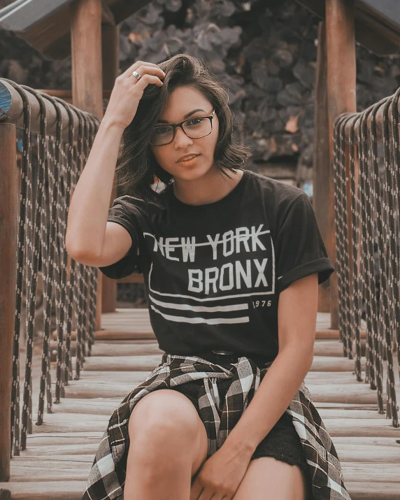 a woman sitting on a bridge wearing a new york bronx t - shirt, by Robbie Trevino, trending on unsplash, wearing square glasses, lesbians, low quality photo, promo image