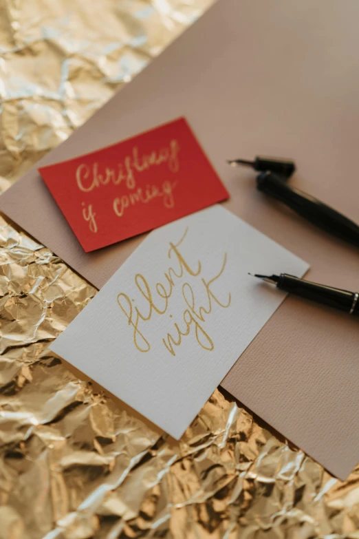 a pen sitting on top of a piece of paper, an album cover, by Julia Pishtar, pexels, christmas night, pair of keycards on table, “modern calligraphy art, red and gold