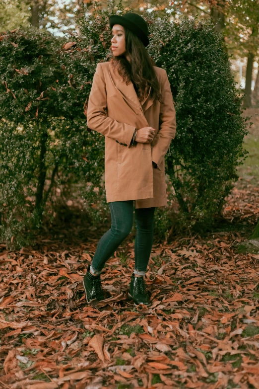 a woman standing in the leaves in a park, inspired by Emma Ríos, trending on pexels, light brown coat, boots, with soft bushes, velvet