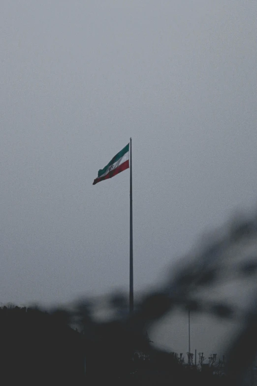 a flag flying on top of a flag pole, a picture, by Attila Meszlenyi, iran, under a gray foggy sky, detention centre, telephoto