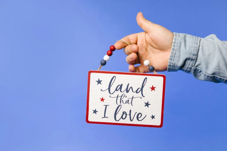a hand holding a sign that says land that i love, decorations, red blue theme, full product shot, proud looking away