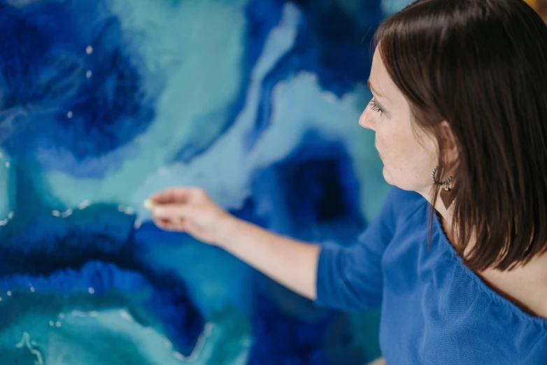 a woman that is standing in front of a painting, by Arabella Rankin, pexels contest winner, process art, blue paint on top, close - up photograph, sapphire waters below, in her art room
