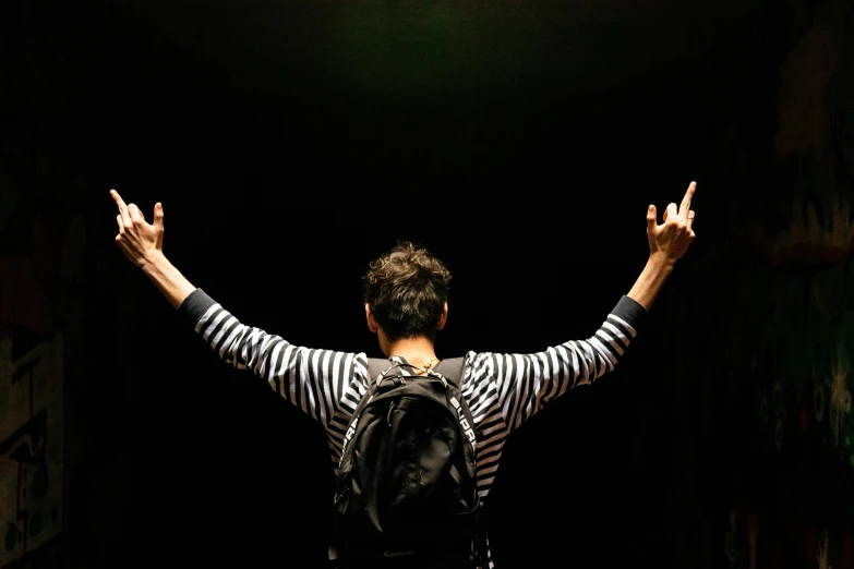 a person standing in a dark room with their hands in the air, by Nina Hamnett, happening, with a backpack, rock star, mime, back view. nuri iyem
