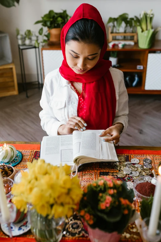 a woman sitting at a table reading a book, hurufiyya, holy themed, books and flowers, head scarf, on a table