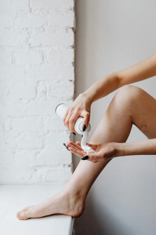 a woman sitting on a window sill holding a bottle of lotion, by Adam Marczyński, trending on pexels, renaissance, shows a leg, flesh + technology, on a white table, shaved