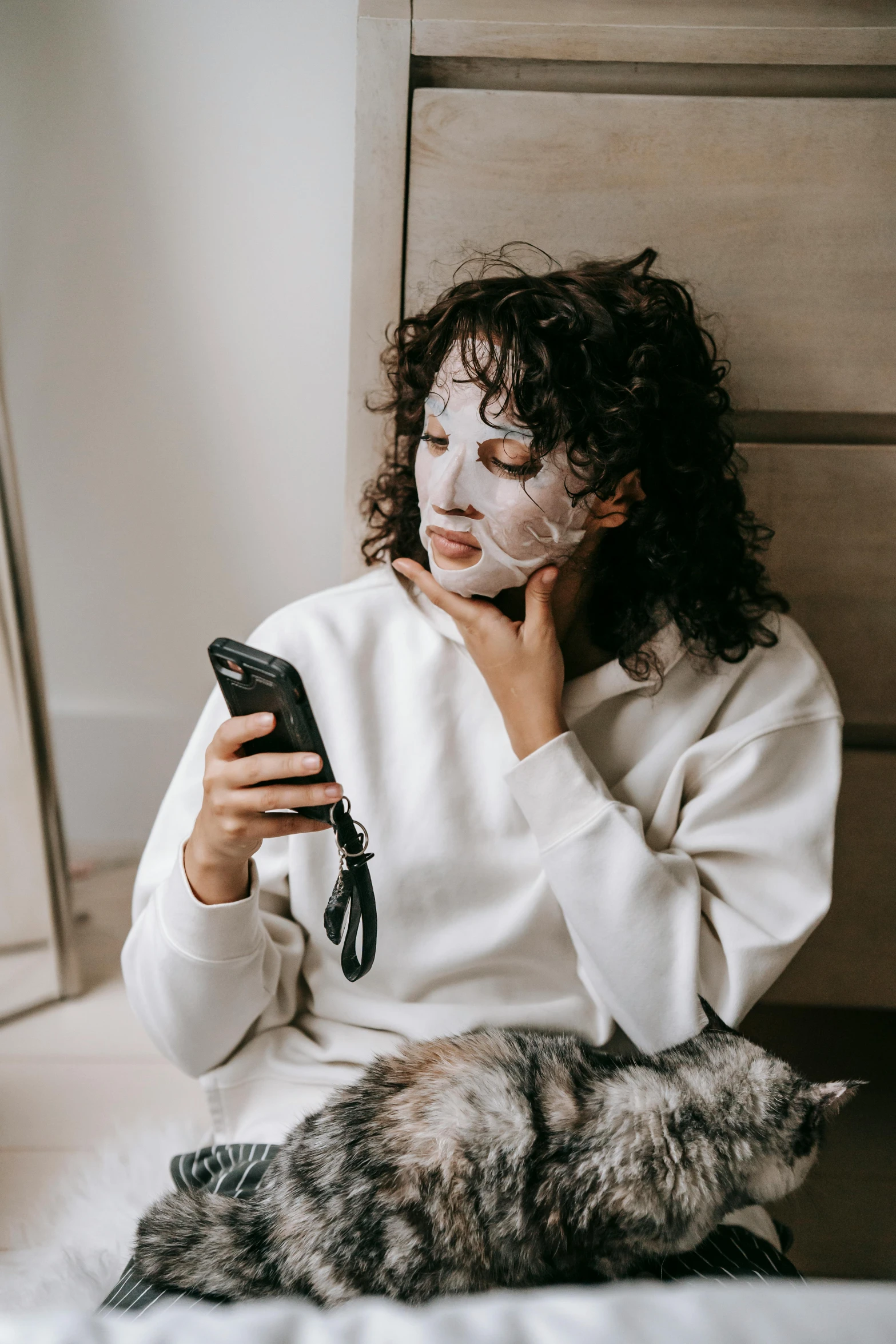 a woman sitting on top of a bed next to a cat, trending on pexels, happening, geisha mask, cellphone, her face is coated in a white, wearing sweatshirt
