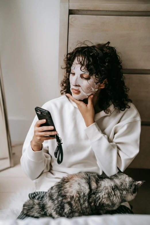 a woman sitting on top of a bed next to a cat, trending on pexels, happening, geisha mask, cellphone, her face is coated in a white, wearing sweatshirt