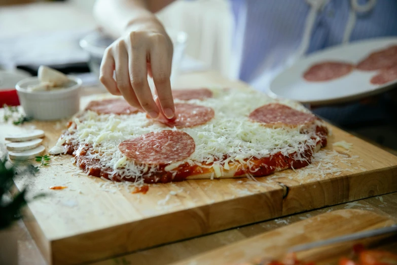 a person making a pizza on a cutting board, looking towards the camera, cheese and salami on the table, no crop, 🦩🪐🐞👩🏻🦳
