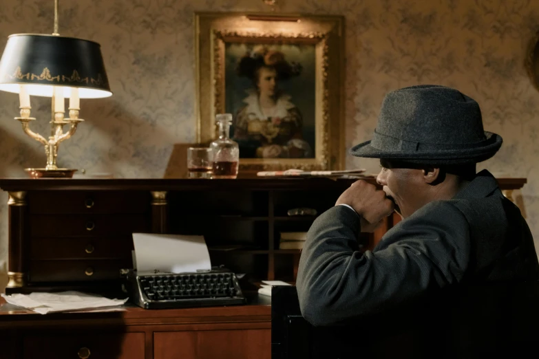 a man sitting at a desk with a typewriter, inspired by Sir William Orpen, pexels contest winner, harlem renaissance, full scene shot, mystery and detective themed, caracter with brown hat, looking at monitor