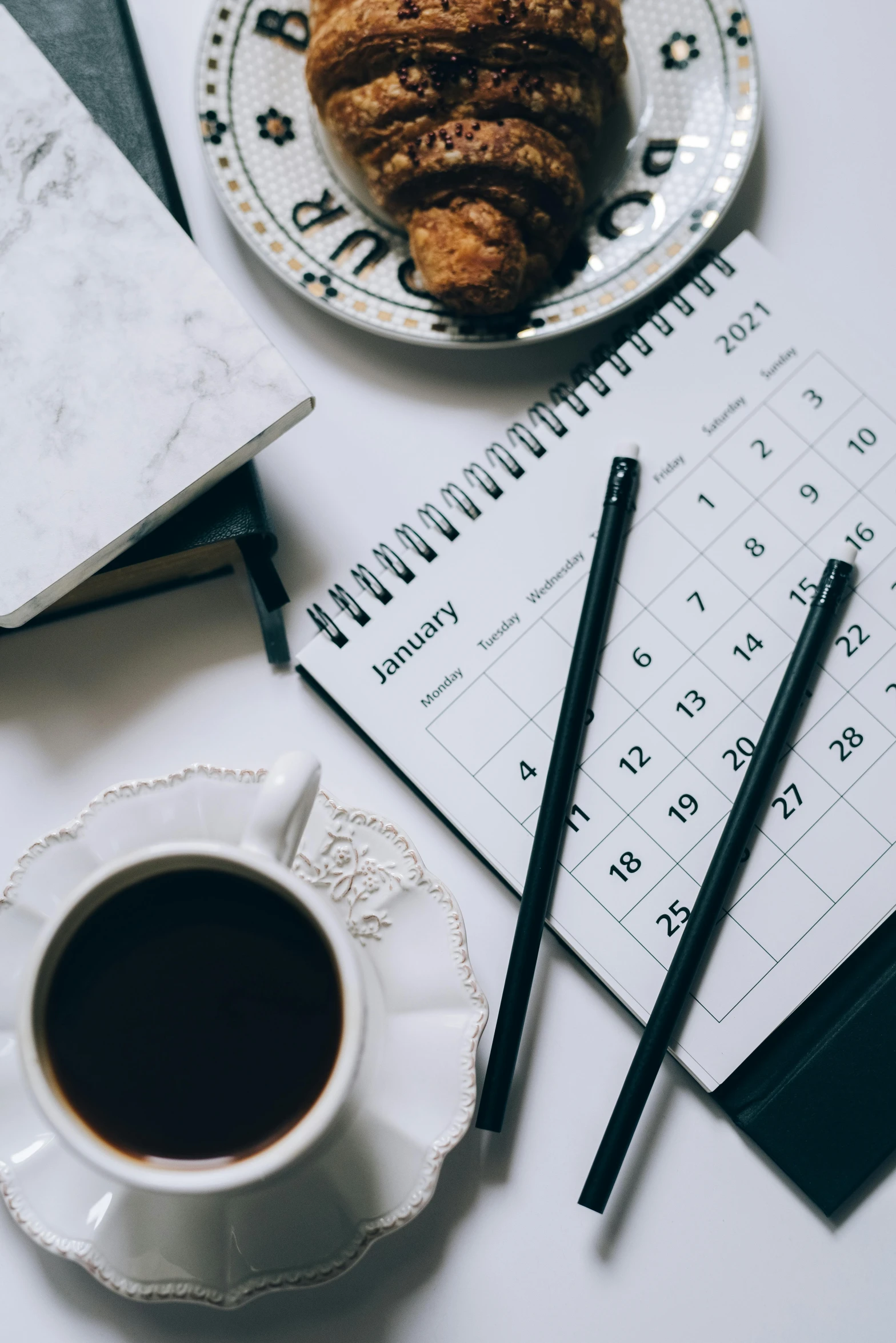 a white plate topped with a croissant next to a cup of coffee, trending on unsplash, happening, female calendar, black ball pen on white paper, thumbnail, cubes on table