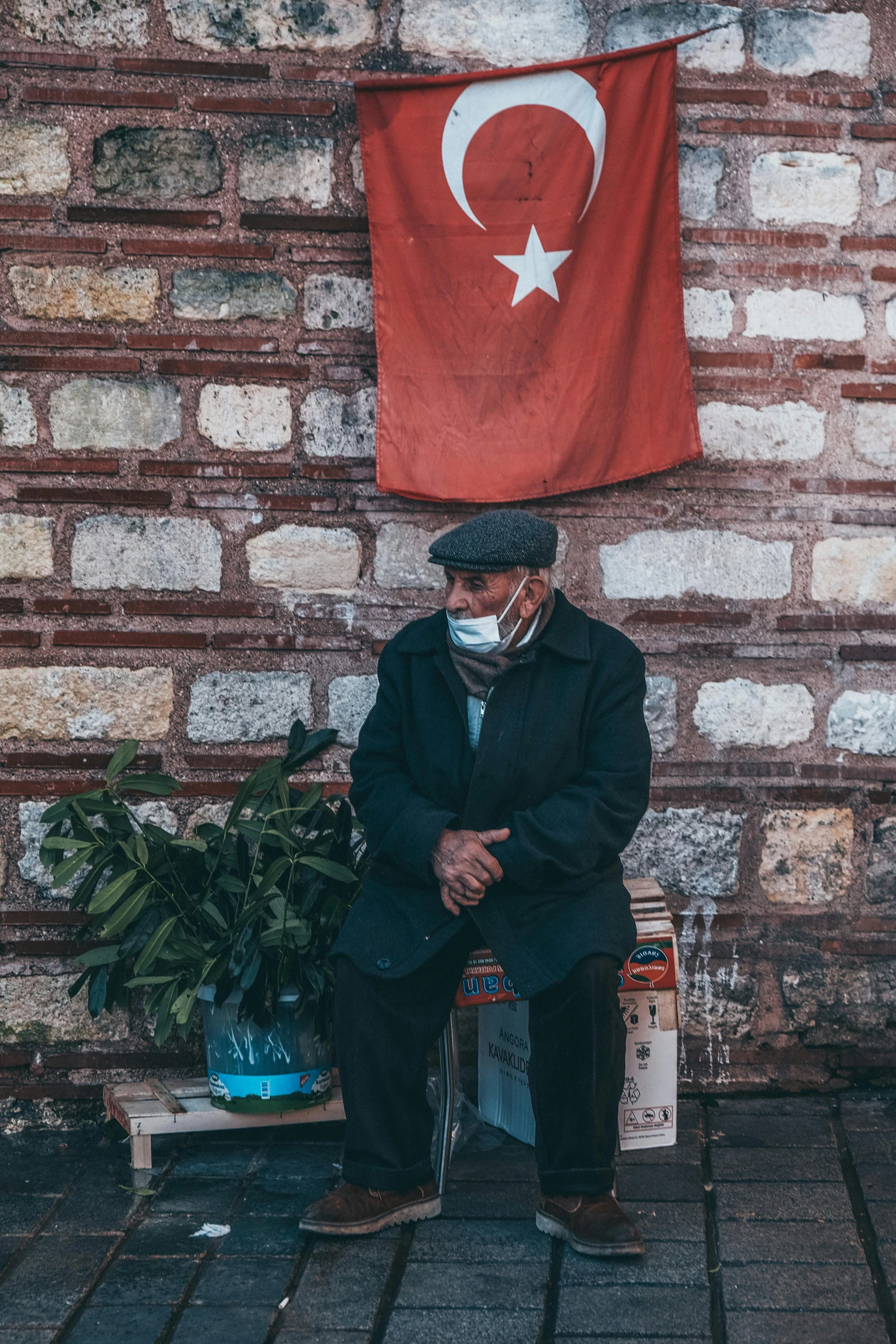 a man sitting in front of a wall with a turkey flag on it, pexels contest winner, symbolism, old man doing with mask, market setting, square, next to a plant
