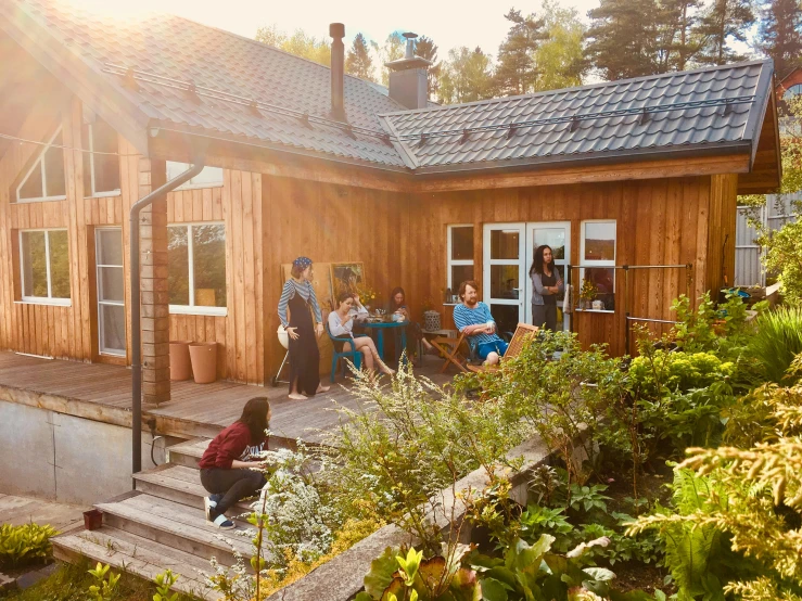 a group of people standing outside of a wooden house, by Jaakko Mattila, pexels contest winner, plants and patio, sun overhead, sitting in the garden, warm coloured