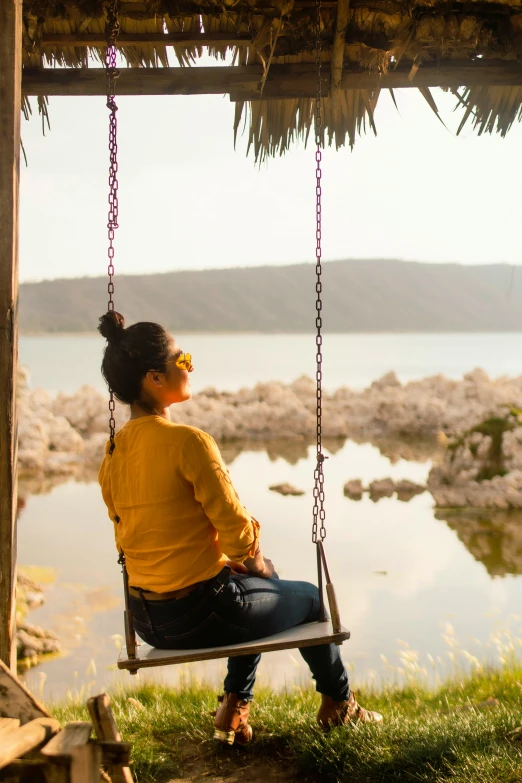 a woman sitting on a swing next to a body of water, pondering, assam tea garden setting, uncropped, yellow