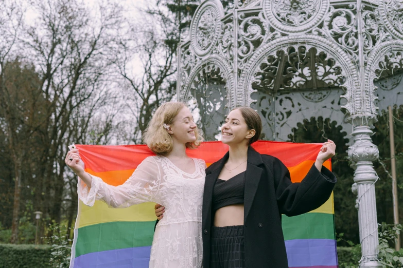 two women holding a rainbow flag in front of a gazebo, trending on pexels, marat zakirov, holding a white flag, 🚿🗝📝