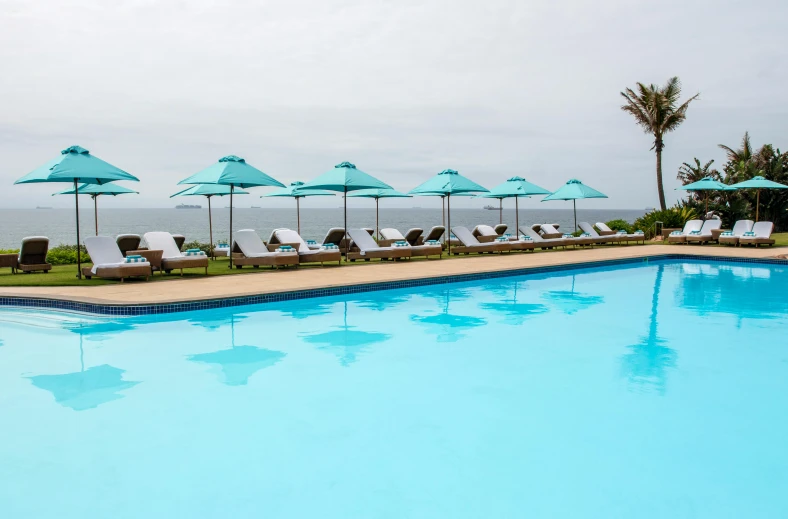 a pool with lounge chairs and umbrellas next to the ocean, teal suit, south african coast, profile pic, cerulean