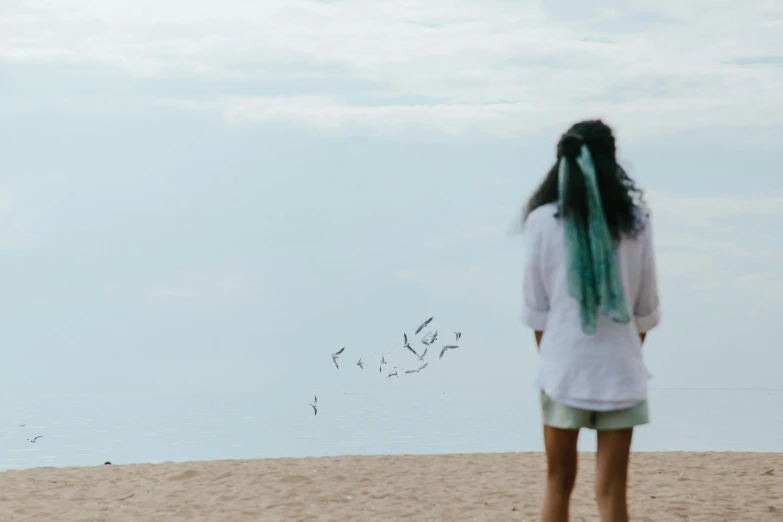 a woman standing on a beach flying a kite, an album cover, by Lucia Peka, pexels contest winner, birds flying in the distance, teal aesthetic, unfocused, facing away