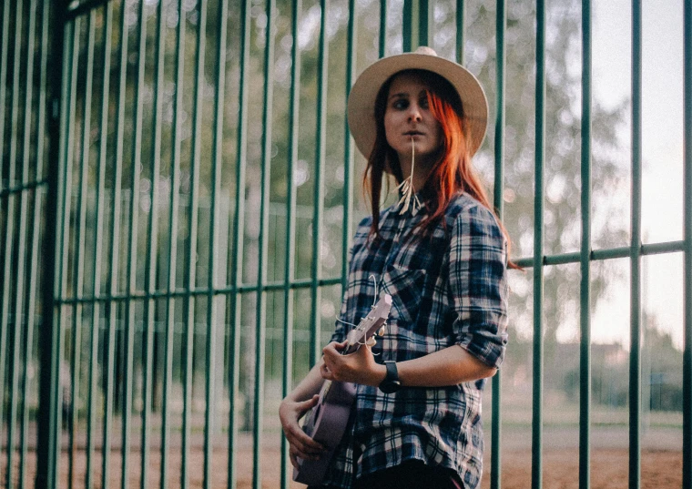 a woman wearing a hat standing in front of a fence, by Julia Pishtar, pexels contest winner, carrying a guitar, redhead girl, plaid shirt, at the park