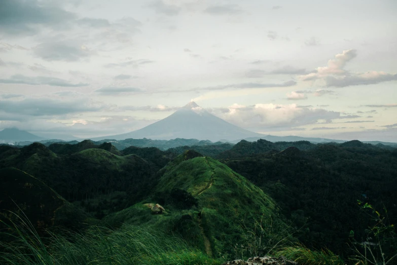 a man standing on top of a lush green hillside, a picture, unsplash contest winner, sumatraism, with a volcano in the background, seen from afar, screensaver, grey