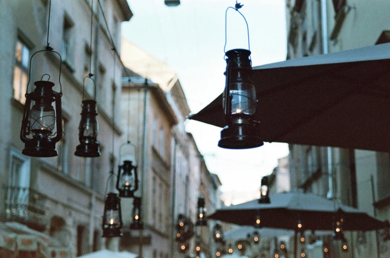 a group of people walking down a street next to tall buildings, by Emma Andijewska, unsplash, photorealism, lanterns on the porch, one single gas lamp, vienna, cafe lighting