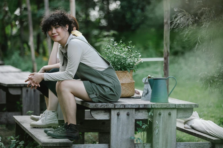 a woman sitting on a bench next to a potted plant, wearing overalls, casual green clothing, charli bowater and artgeem, on a wooden table