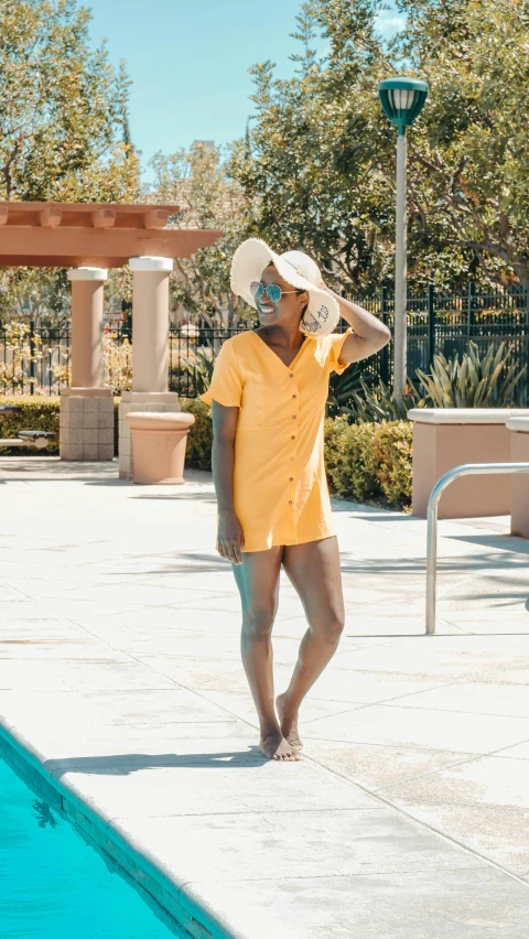 a woman standing next to a swimming pool, yellow overall, tan skin a tee shirt and shorts, thumbnail, sleek robes