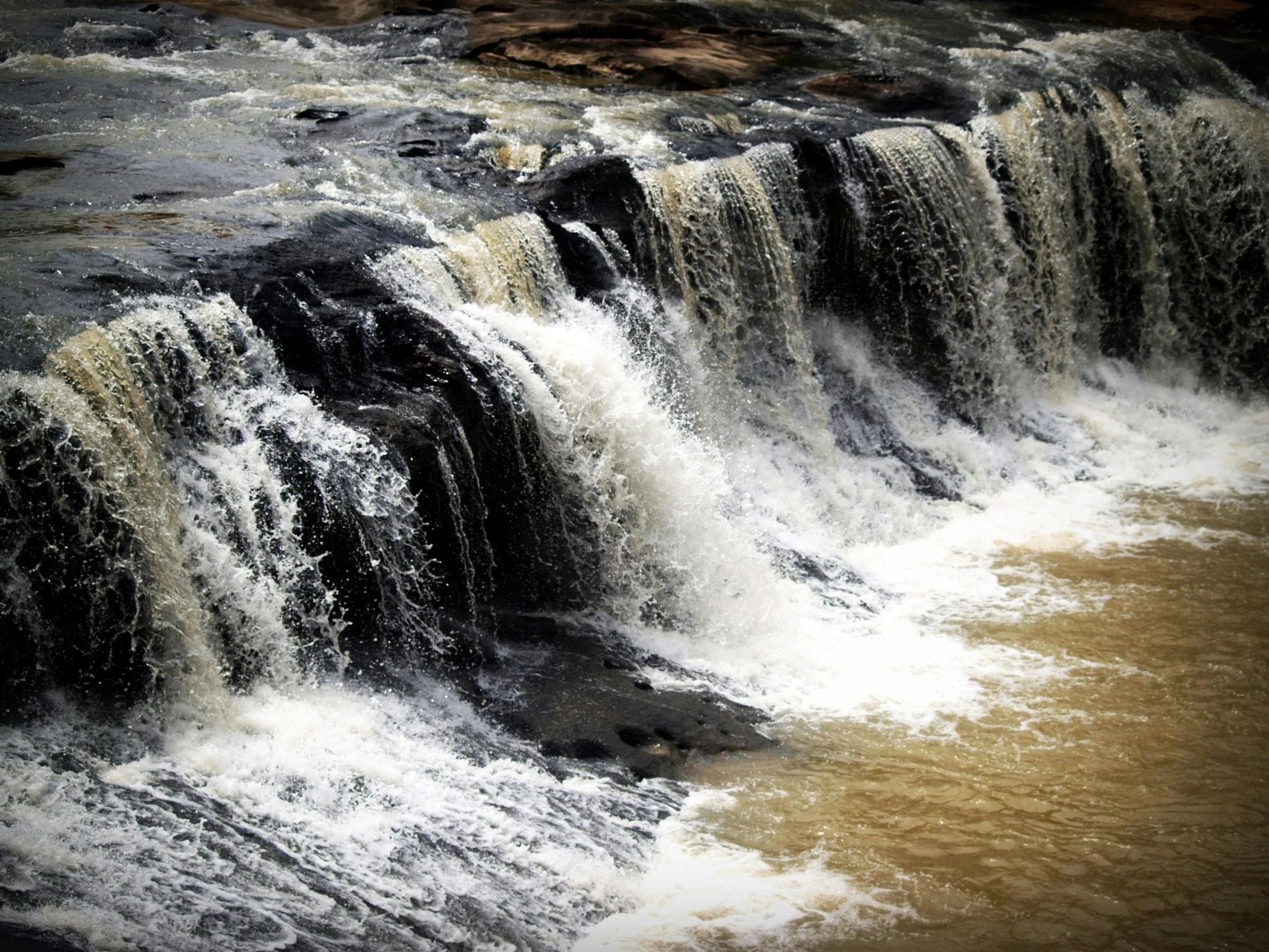 a large waterfall in the middle of a river, pexels contest winner, hurufiyya, dark yellowish water, thumbnail, sandfalls, black