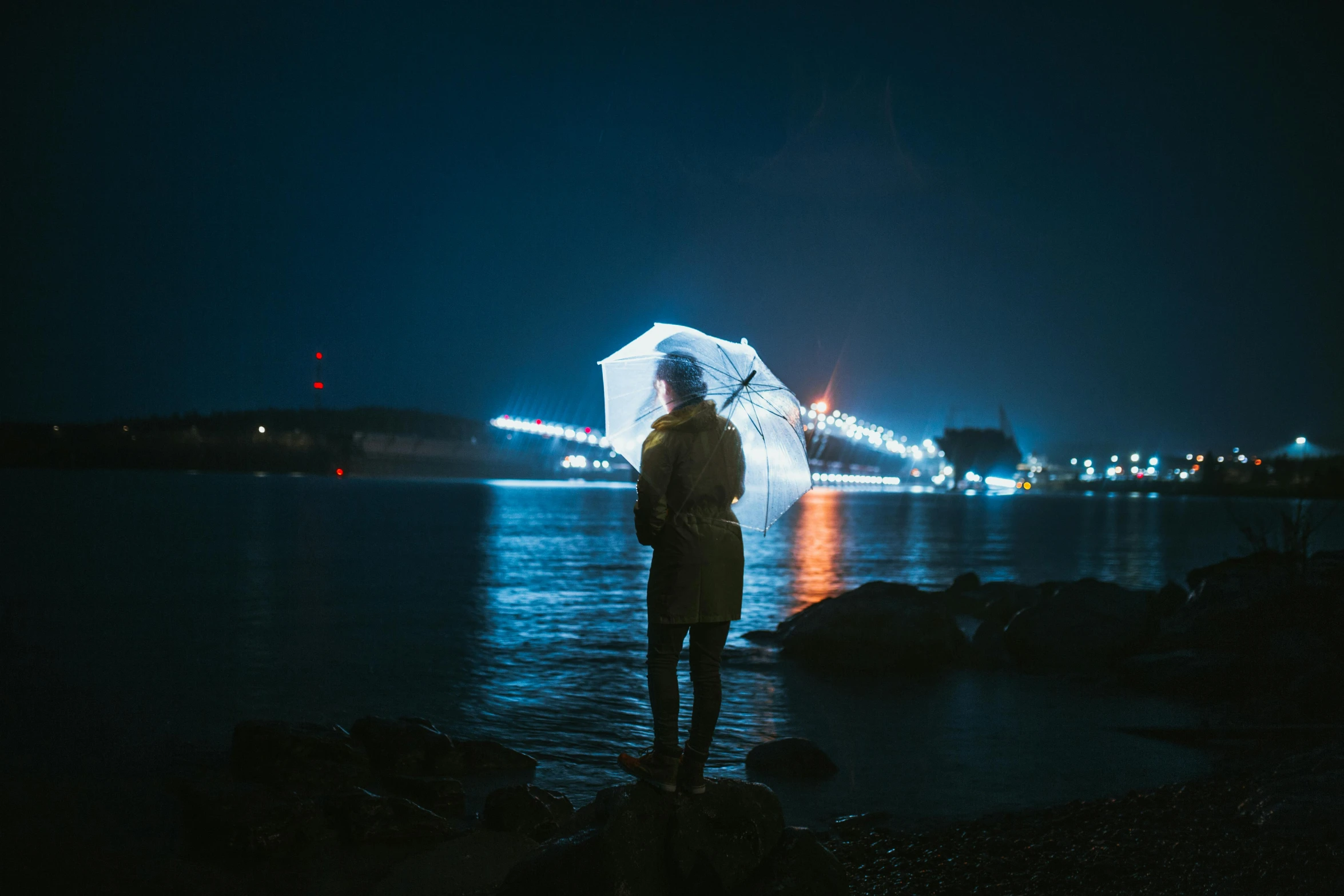 a person standing in front of a body of water holding an umbrella, inspired by Elsa Bleda, unsplash contest winner, dramatic white and blue lighting, night lights, backlit shot girl in parka, instagram post