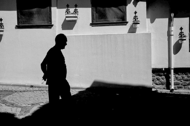 a black and white photo of a man walking down a street, a black and white photo, by Ferenc Joachim, minimalism, deep shadows and colors, silhouette!!!, in front of the house, ludek pesek