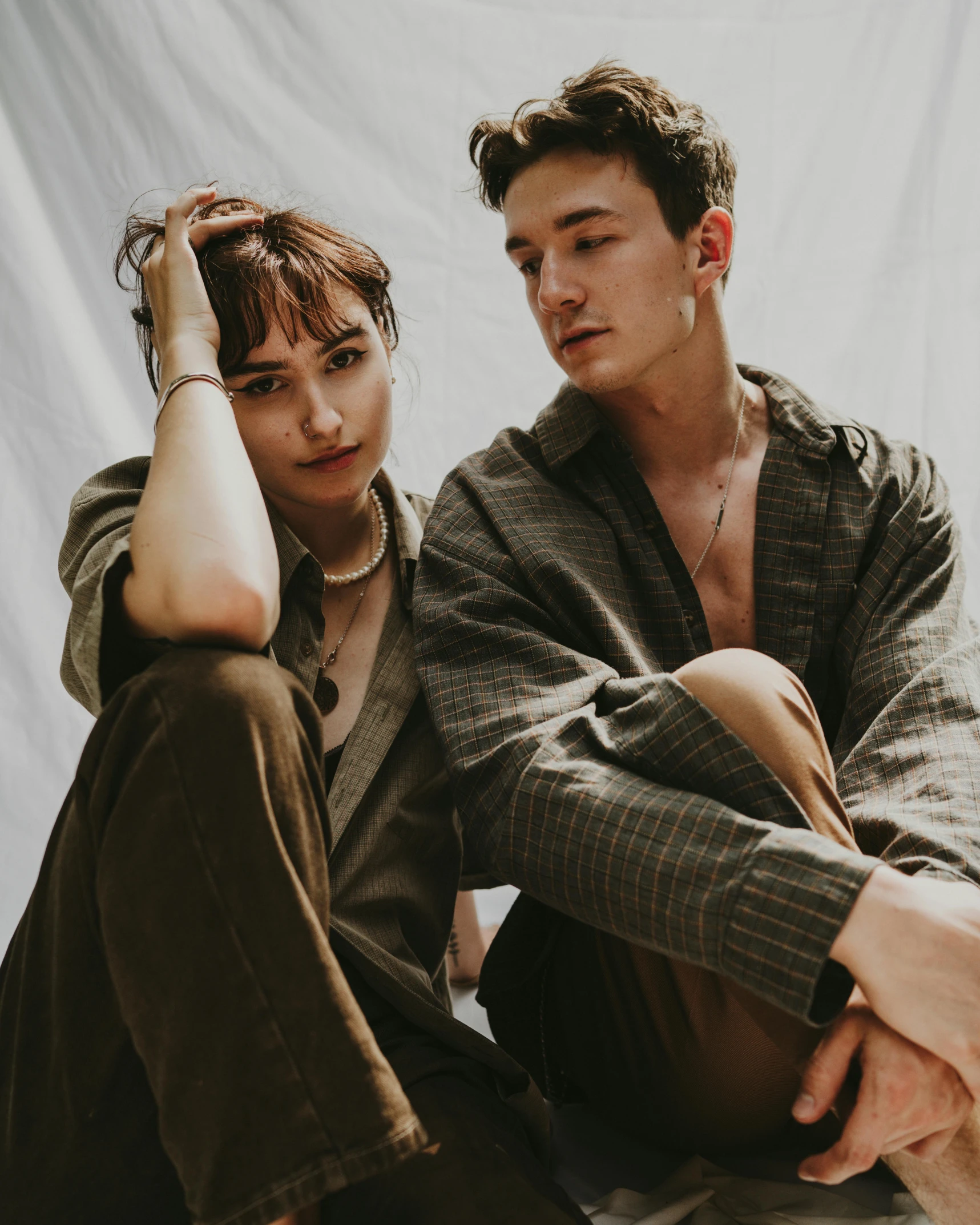 a man and a woman sitting next to each other, an album cover, trending on pexels, beautiful androgynous girl, lily frank, brown clothes, proud looking away