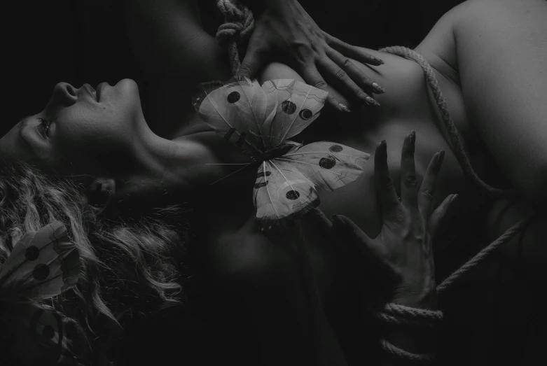 a woman laying on top of a bed next to a butterfly, a black and white photo, by Adam Marczyński, pexels contest winner, surrealism, shibari, woman holding another woman, in the dark, a close up shot