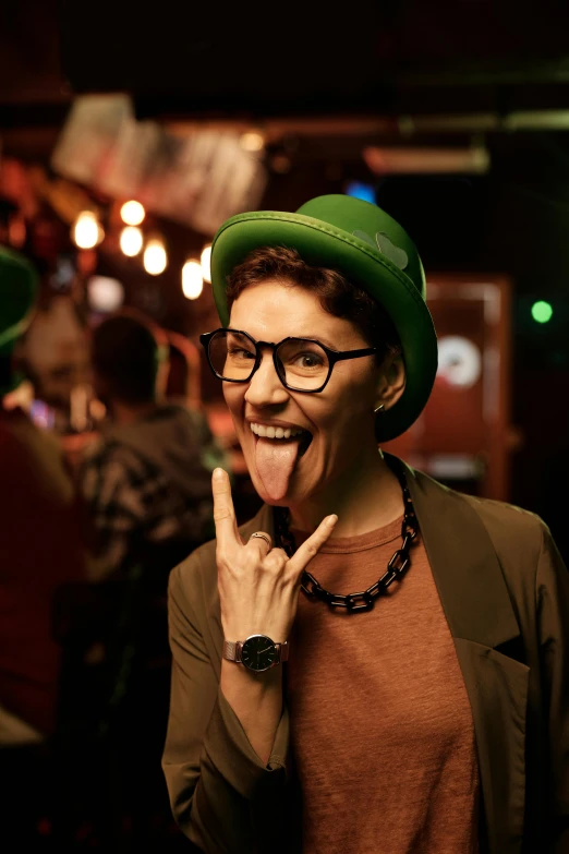 a woman with glasses and a green hat making a peace sign, trending on pexels, happening, in a pub, licking tongue, androgyny, college party