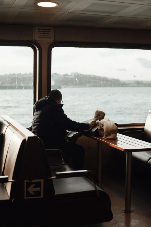 a man sitting at a table in front of a window, on a boat, 🚿🗝📝, overcast weather, coffee smell