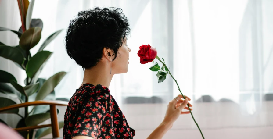 a woman sitting at a table with a flower in her hand, pexels contest winner, romanticism, profile image, holding a red rose, androgynous person, looking across the shoulder