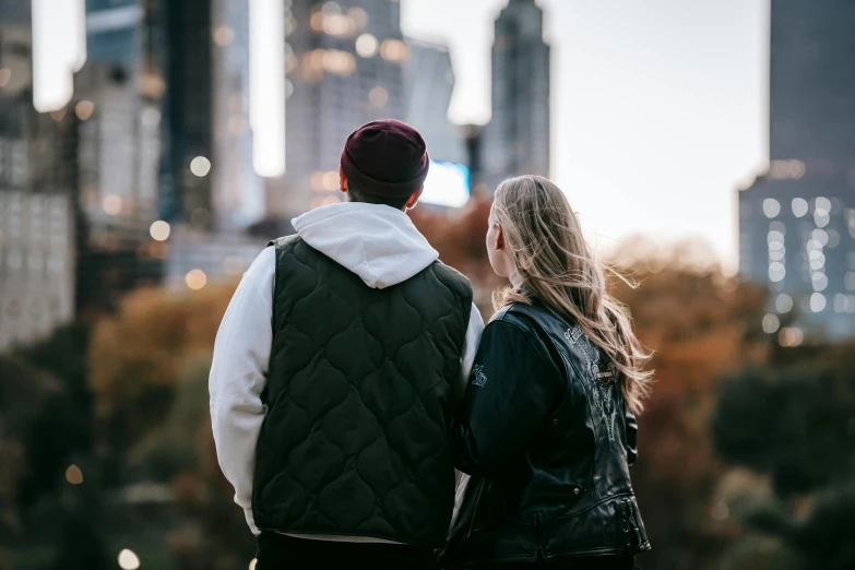 a man and a woman standing in front of a city skyline, trending on pexels, park in background, looking from behind, leather clothing, ayne haag