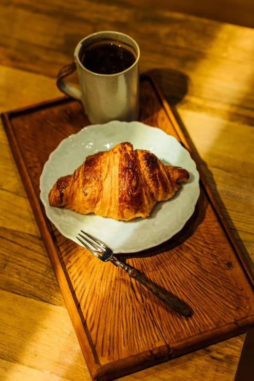 a white plate topped with a croissant next to a cup of coffee, a portrait, by Sven Erixson, unsplash, on a wooden tray, directional sunlight skewed shot, jen atkin, cone shaped