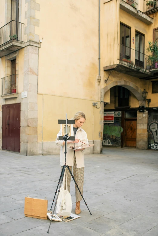 a woman standing next to a camera on a tripod, an album cover, inspired by Modest Urgell, plein air, gothic quarter, slight yellow hue, stands at a his easel, streetview