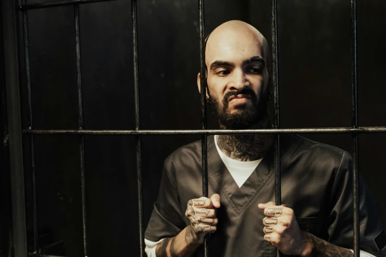 a man standing behind bars in a jail cell, a portrait, inspired by James Bolivar Manson, pexels contest winner, hurufiyya, tattooed, wearing prison jumpsuit, nipsey hussle, bald