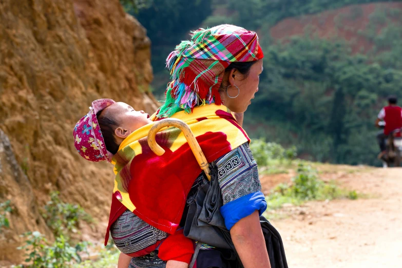 a woman carrying a child on her back, by Alice Mason, pexels contest winner, cloisonnism, wearing a red backwards cap, vietnam, multicoloured, holding an epée