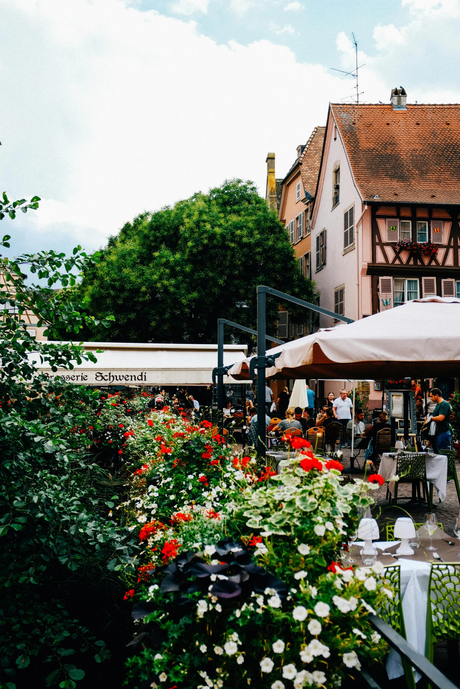 a group of people sitting under umbrellas in front of a building, inspired by August Querfurt, unsplash, renaissance, rich vines and verdant flowers, cafe tables, white buildings with red roofs, marketplace