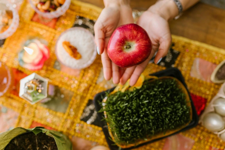 a person holding an apple on top of a table, by Julia Pishtar, trending on pexels, ayahuasca ceremony, the table is full of food, square, hindu aesthetic